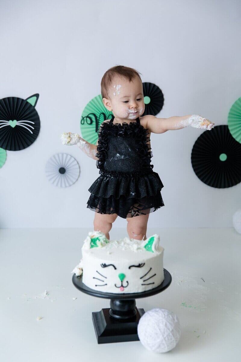 A baby in a black frilly outfit stands on a table covered in frosting, with a cat-themed cake in front.