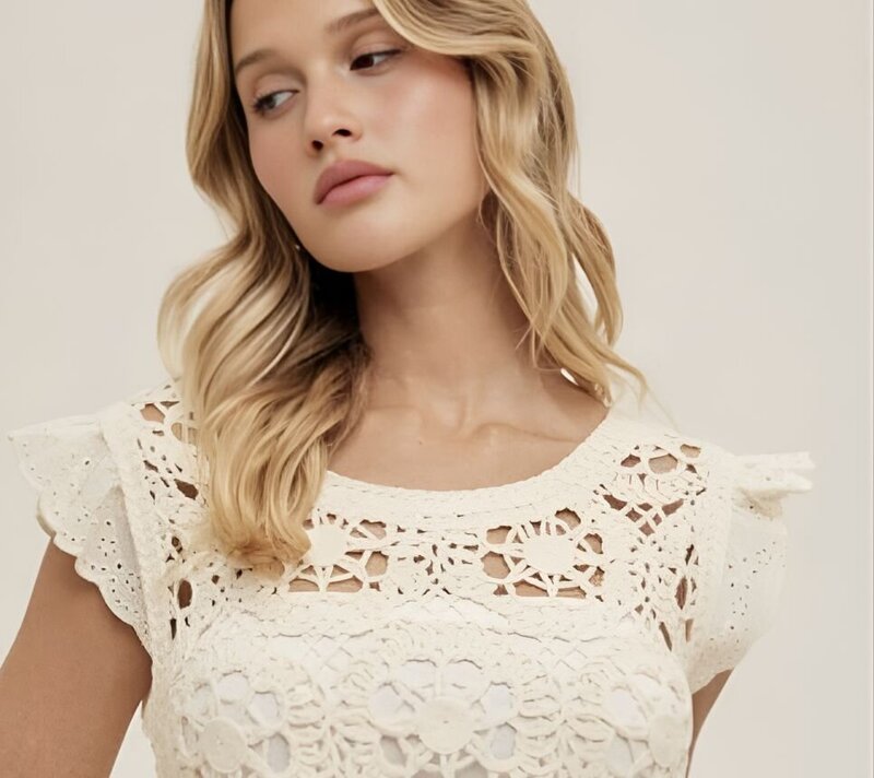 An image of a young blonde woman in her thirties with slightly curled hair wearing a stylish off-white crochet top with a flower pattern.
