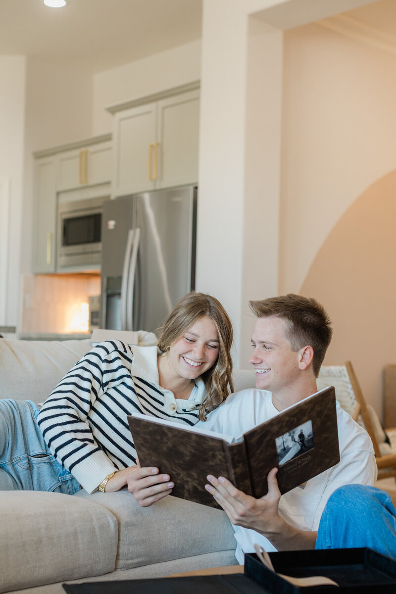 A joyful couple relaxes on a cozy beige couch in their modern home, flipping through their elegant wedding album filled with cherished memories. Captured by an Alabama wedding photography expert, this moment highlights the beauty of preserving love stories through timeless imagery.