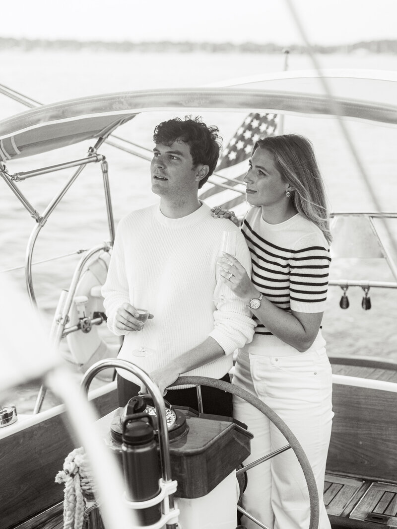 A black and white photo of a couple steering a sailboat at their Newport engagement photo session.