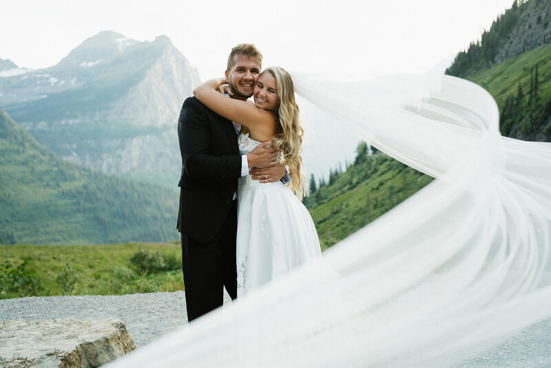 Elopement or wedding photos taken in Glacier National Park. The bride is hugging the groom with her arms around his neck and his arms around her waist. They are smiling at the camera and her veil reaches the camera with the wind blowing it magestically.