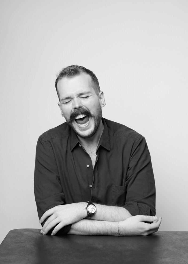 Black and white portrait of a man mid-laugh, eyes closed and mouth open in an uninhibited moment of joy. He wears a relaxed button-up shirt, accessorized with a watch and bracelet, his arms casually crossed on the table. A bold, unfiltered moment from 'THIS IS ME – A Photography Experience by Katie Campbell' in Atlanta, GA, celebrating personality and authentic self-expression.