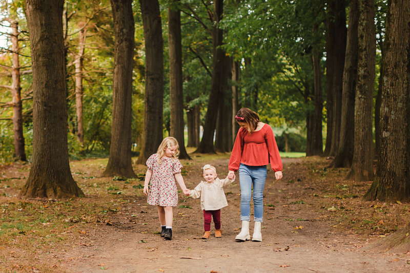 Three young girls holding hands walk together outside on tree lined path