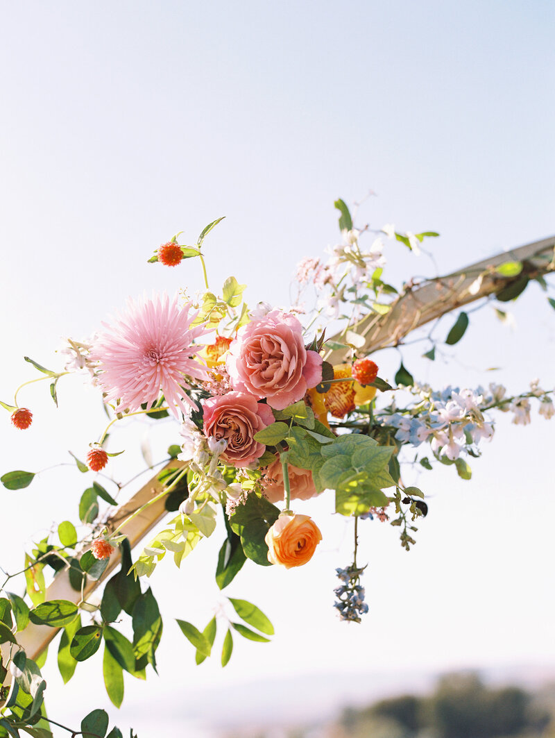 A closeup detail of the ceremony's tropical and bright colored flowers on a gold arch, designed by Moonlight Floral Co. for the wedding at Dos Pueblos Orchid Farm, photographed by My Sun and Stars Co.