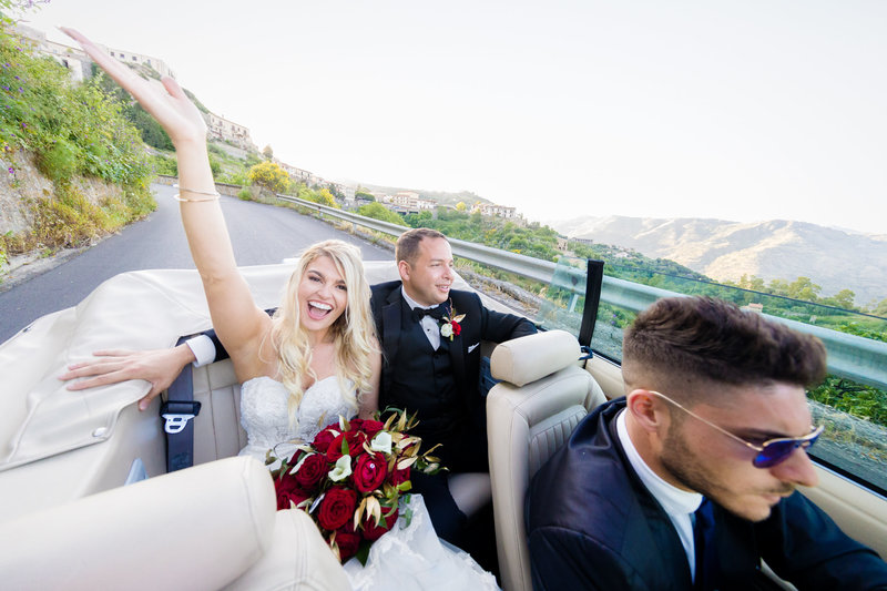 Bride throwing her hand up in joy while riding in the back of a limo in Italy, showcasing celebratory wedding style.
