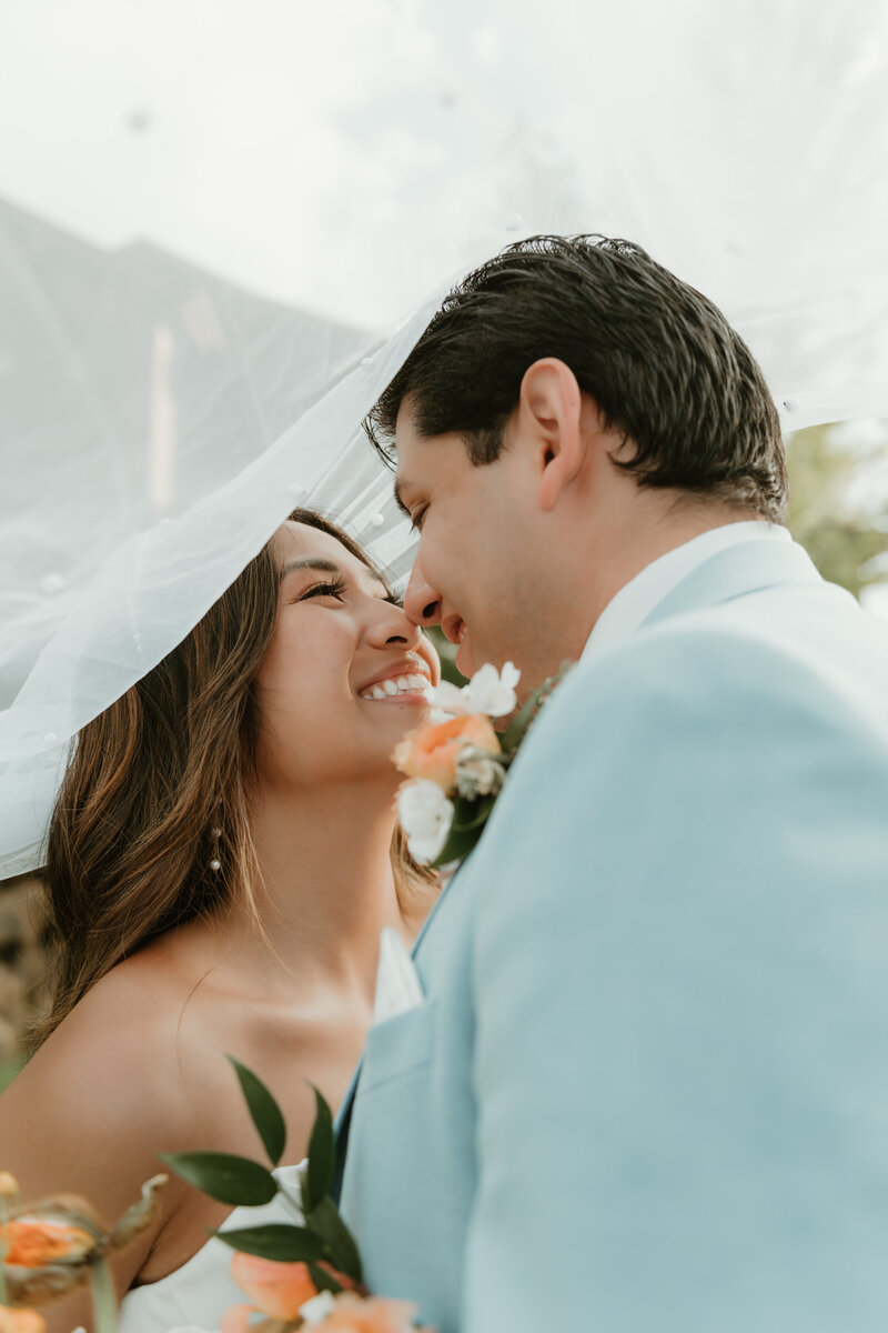 Photo of a bride and groom taken by Bailee Freeman Photography. The photo is a veil shot where the bride and groom are underneath the veil and their faces are close together in an almost kiss. Groom is in a powder blue suit with a peach boutonniere.