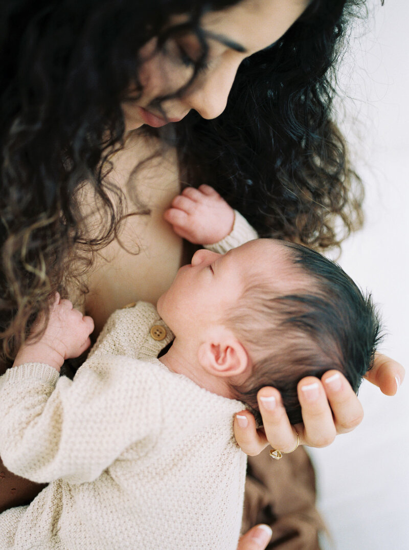 Woman with dark curly hair holds newborn infant child's head in her hands while she lovingly gazes at him while he sleeps and is wearing a beige sweater by Portland Newborn Photographer Emilie Phillipson Photography