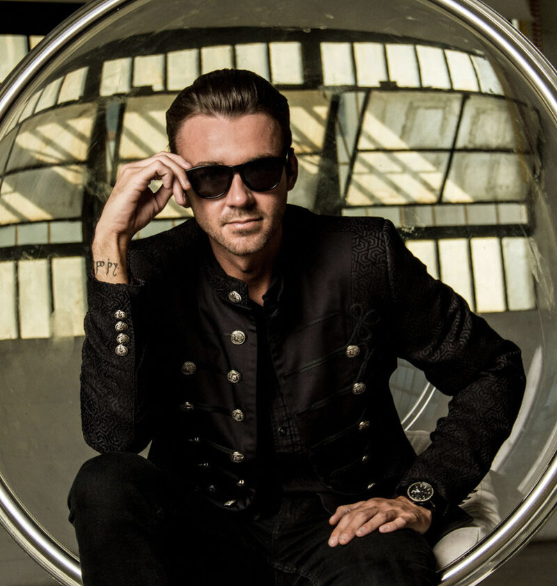 Professional Branding Portrait Author Chandler Morrison sitting in clear circle chain wearing dark suit one hand holding sunglasses  stem he wears other hand resting on knee