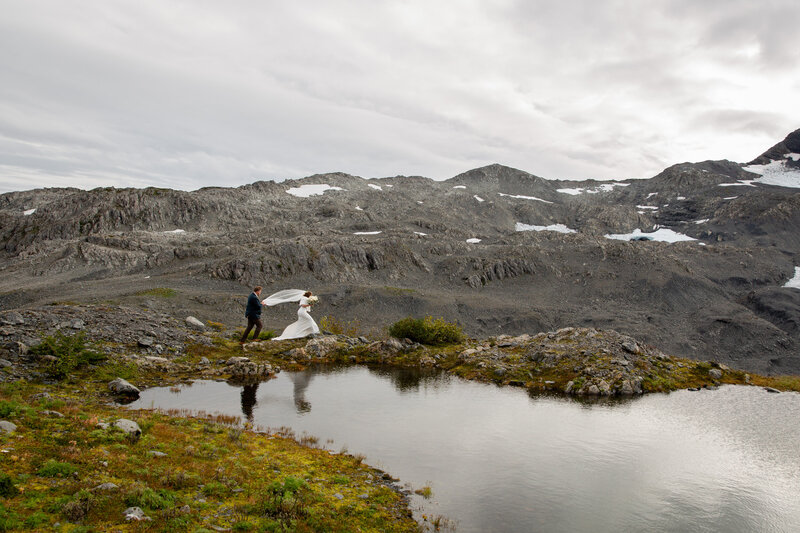 A bride wearing a white dress and a veil walks in front of her groom along the side of a lake in Alaska.