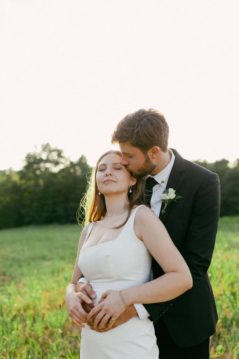 The groom embraces the bride from behind, planting a tender kiss on her cheek during their elopement in the field.