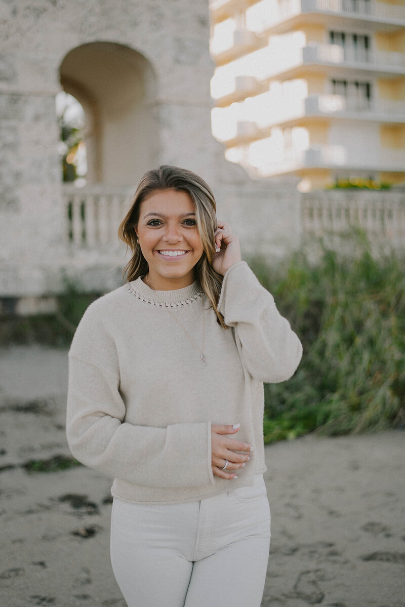 Sleep consultant, Serena Girardi, stands on the sand in a tan sweater and white jeans and smiles