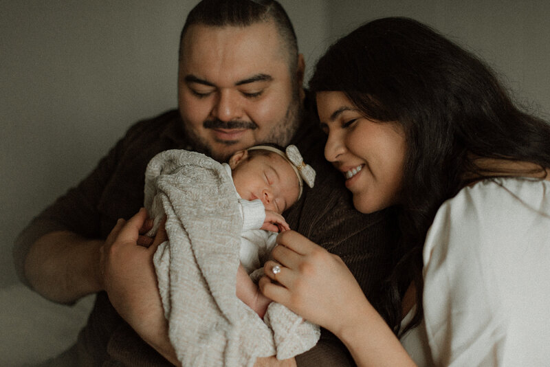 New parents cuddling their swaddled newborn girl, sharing a tender smile in their cozy Murrieta home.