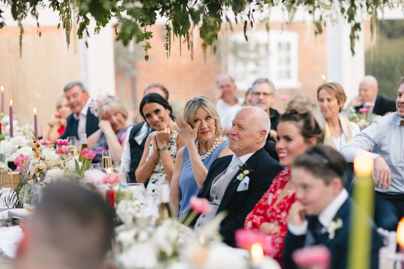 At New Barn Farm, a group of people seated at a long table during wedding speeches adorned with colourful flowers and candles. Some guests are clapping and smiling, while others appear emotional. A canopy draped with hanging greenery adds to the charm of the occasion.