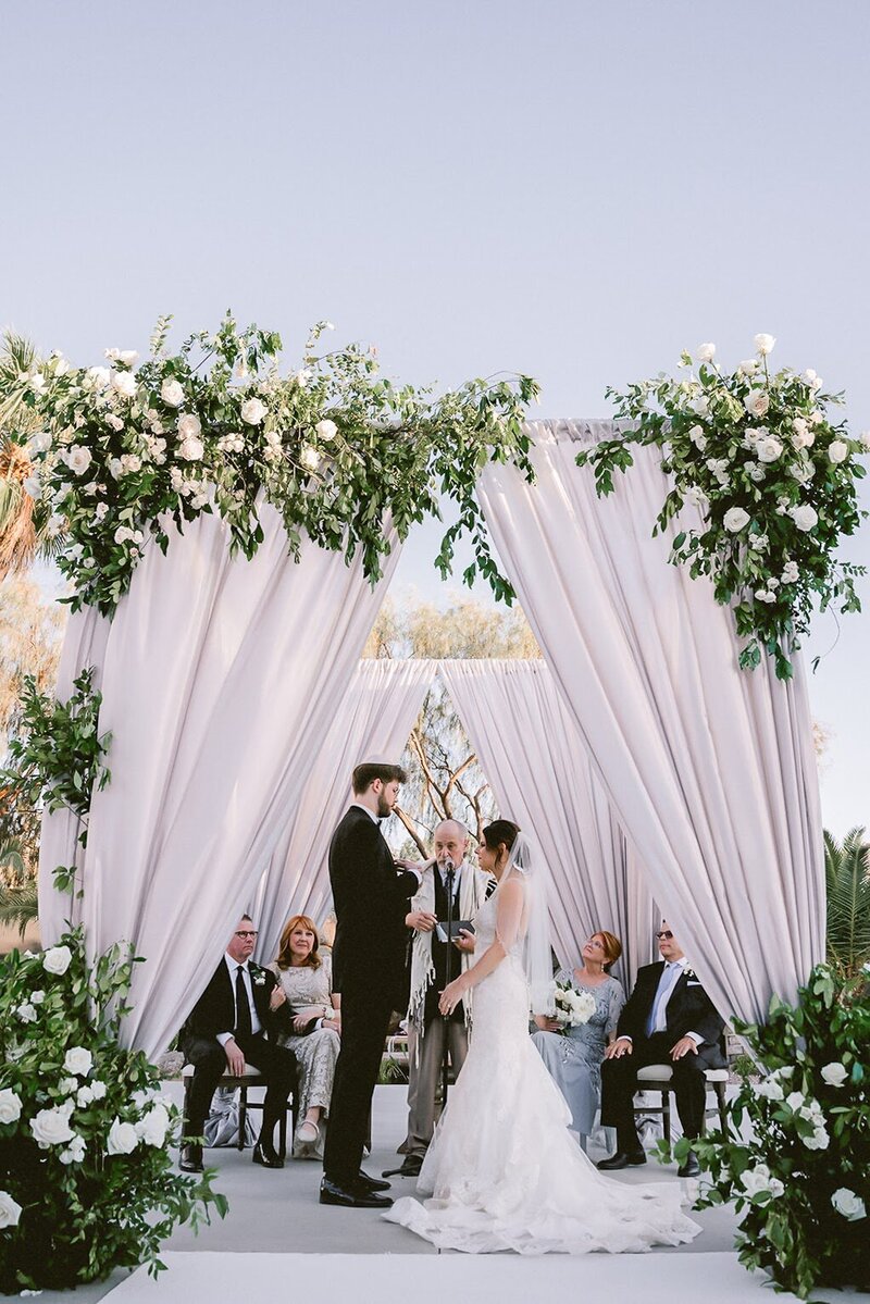 Bride and groom take their wedding vows underneath an elegant draped and floral chuppah