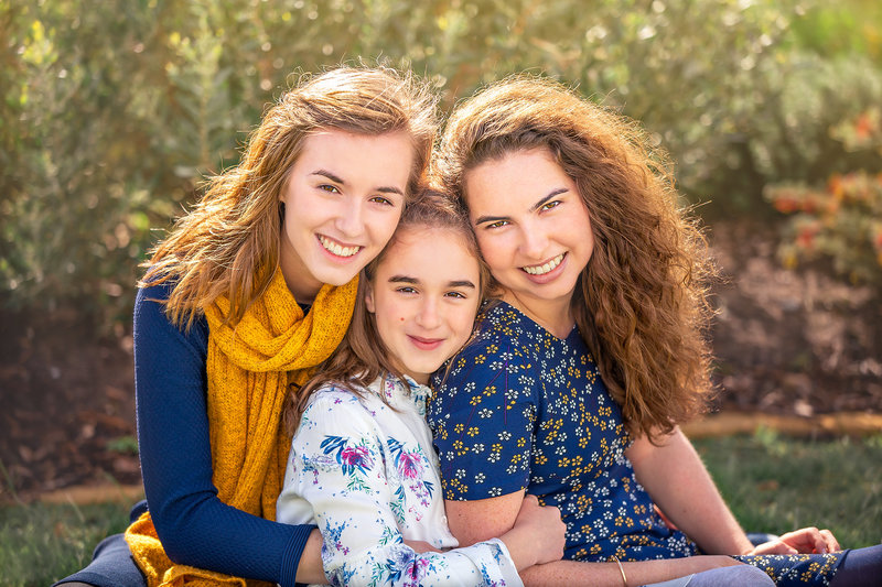 Hampton Roads family photographer captures three sisters sitting together for their outdoor family session hugging each other as the sun shines from behind them