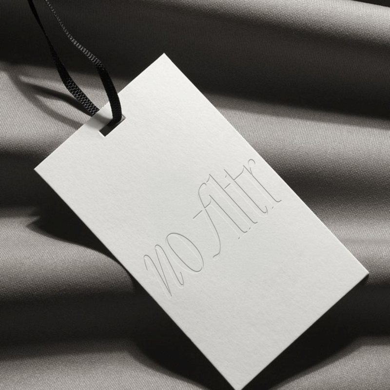 White clothing tag with "No Filtr" in light script, attached by black ribbon on soft gray fabric with subtle folds.