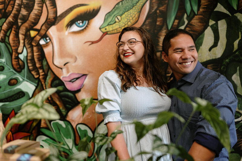 A couple poses closely together amid vibrant tropical foliage and a colorful mural of plants and animals in the background.