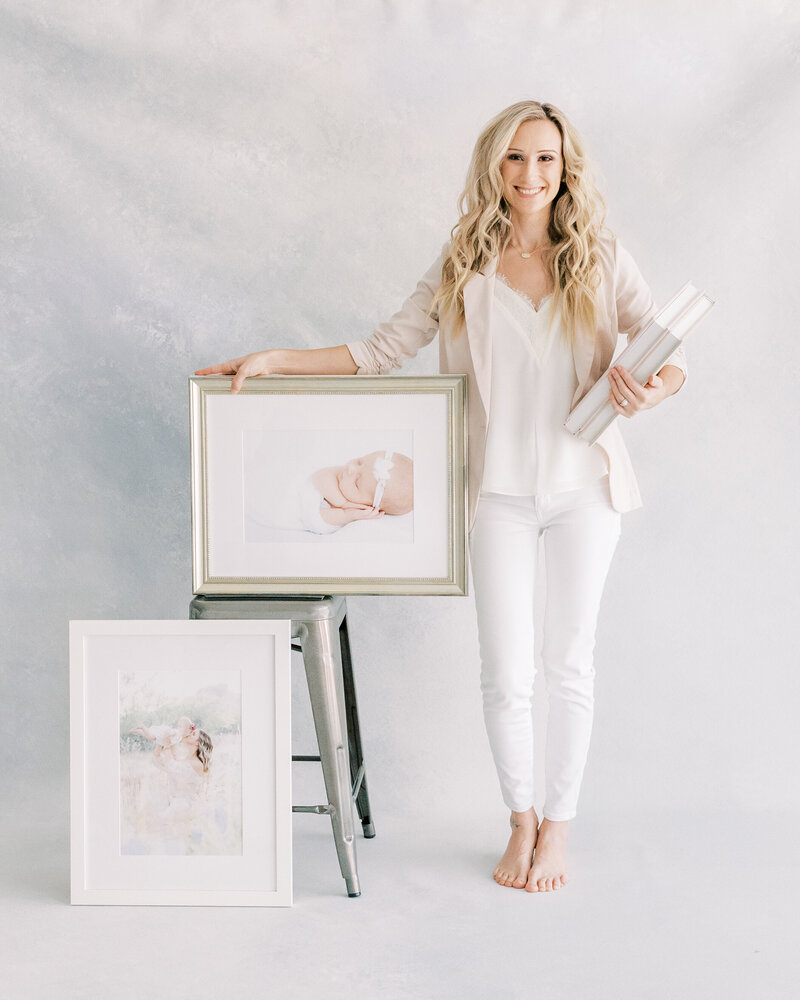 nichole pach standing on a hand painted canvas in her studio holding albums and frames she custom created for her clients in white shirt and blush blazer in Scottsdale Arizona