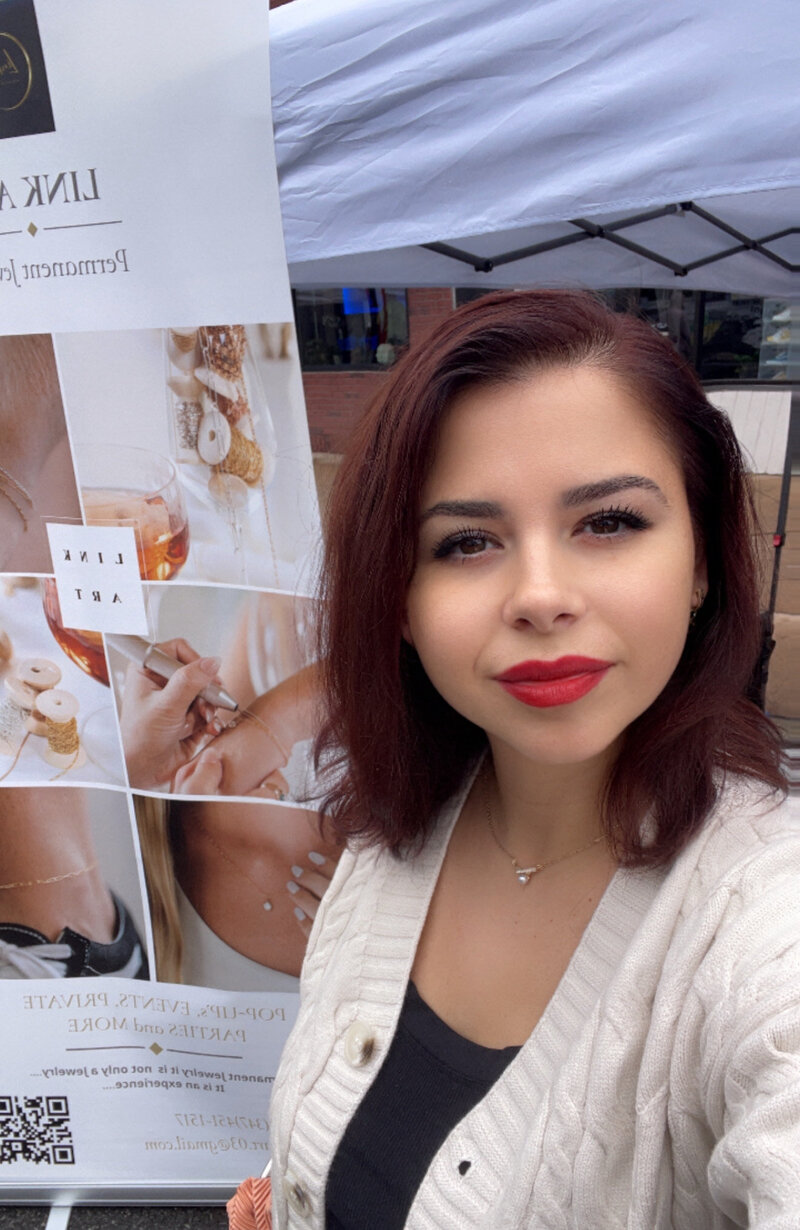 Denisa from Link Art standing in front of her permanent jewelry banner at a pop-up event.