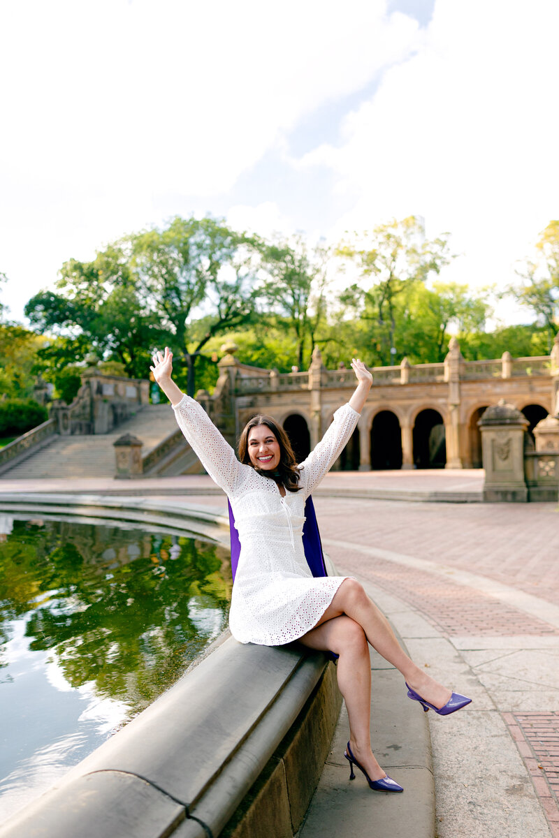NYU graduate celebrating at Bethesda Fountain with her hands raised during a graduation portrait session in Central Park.