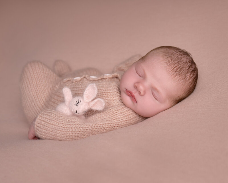 Nyfødt baby i beige strikkedress med liten bamse sover fredelig under fotografering i studio nær Oslo.
