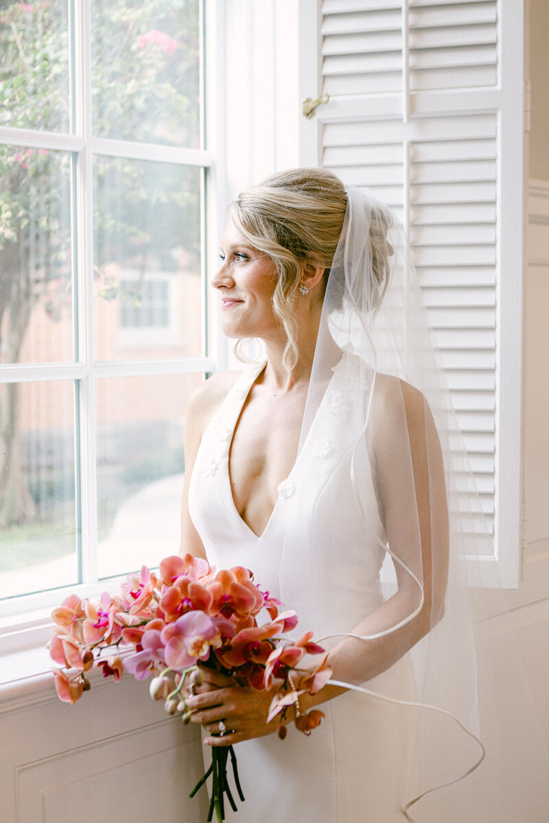 A wedding photo of a bride in a white dress holding her flowers looking out a window