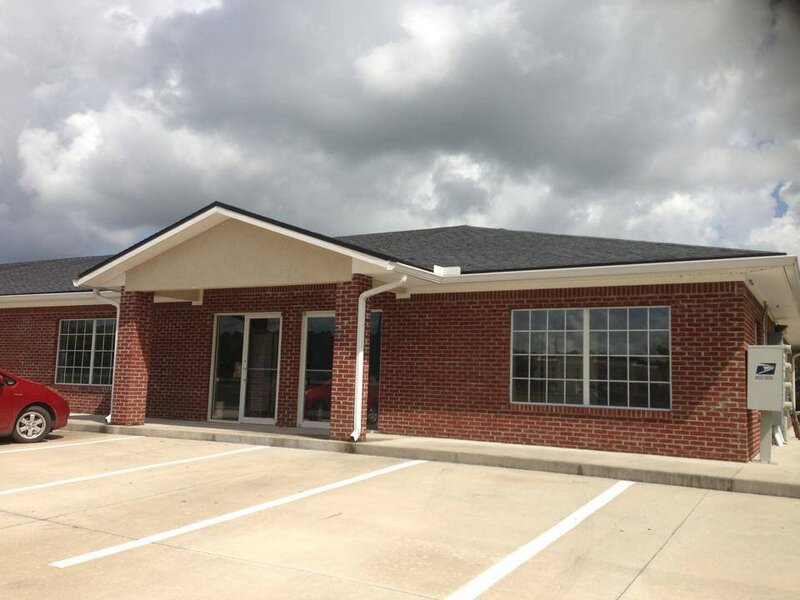 A small, single-story red brick building with a sloped roof, two large windows, and double glass doors stands prominently. The professional photography studio, favored by a Jacksonville headshot photographer, is surrounded by an empty parking lot under a cloudy sky. A red car is partially visible on the left.