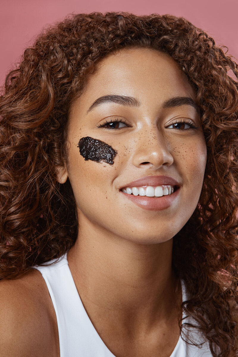 Jayla smiling as she applies a black skincare product to her face, photographed by Ylva Erevall.