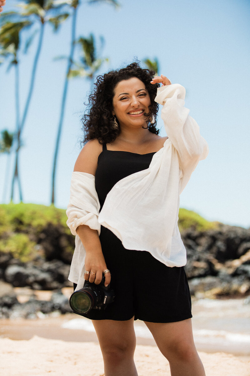 Renata Voci photographed by Nicole Hill on the beaches of Maui, HI. Renata is windswept, tucking her curly hair behind her ear as she holds her camera on the beach with palm trees blurred out behind her int he distance.