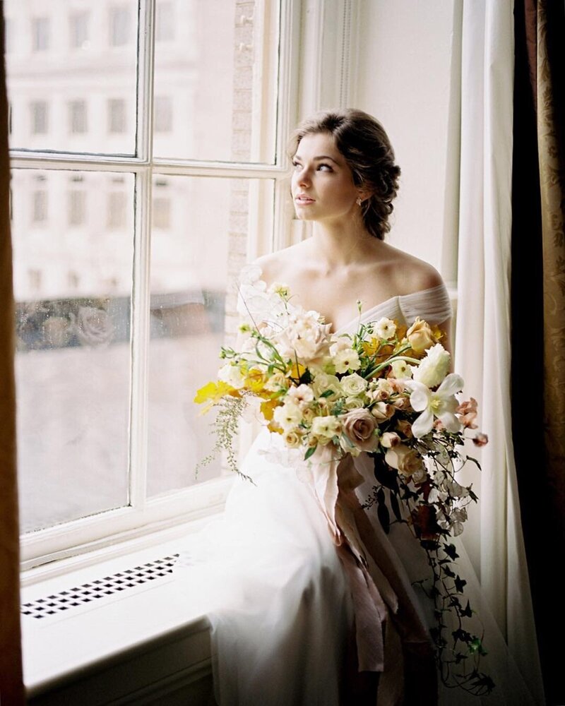 A bride in an off the shoulder dress at a window while a holding bouquet by Pittsburgh Wedding Photographer, Anna Laero.