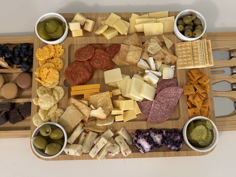 Charcuterie board with a variety of cheeses, cured meats, crackers, olives, pickles, and snacks arranged on a wooden tray, with sweet treats and fruit on the side.