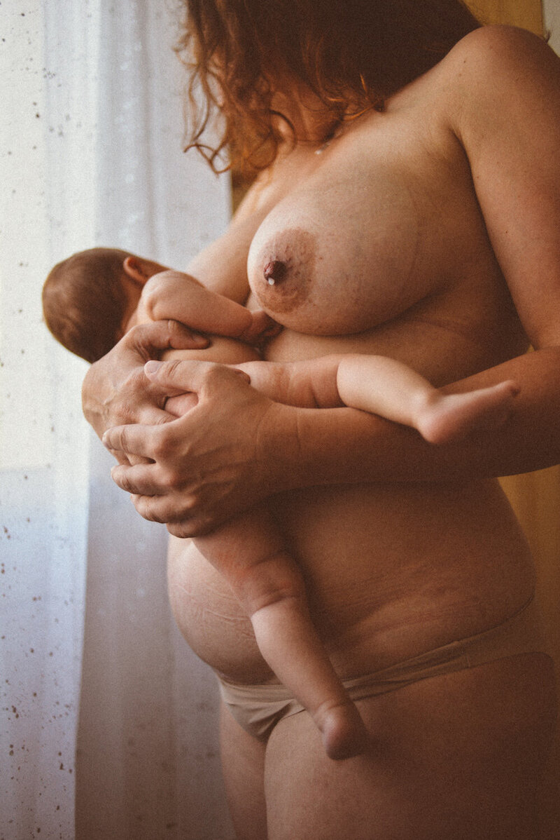 Une femme nue debout qui allaite son bébé tout contre sa peau, une goutte de lait s'échappe de l'autre sein.