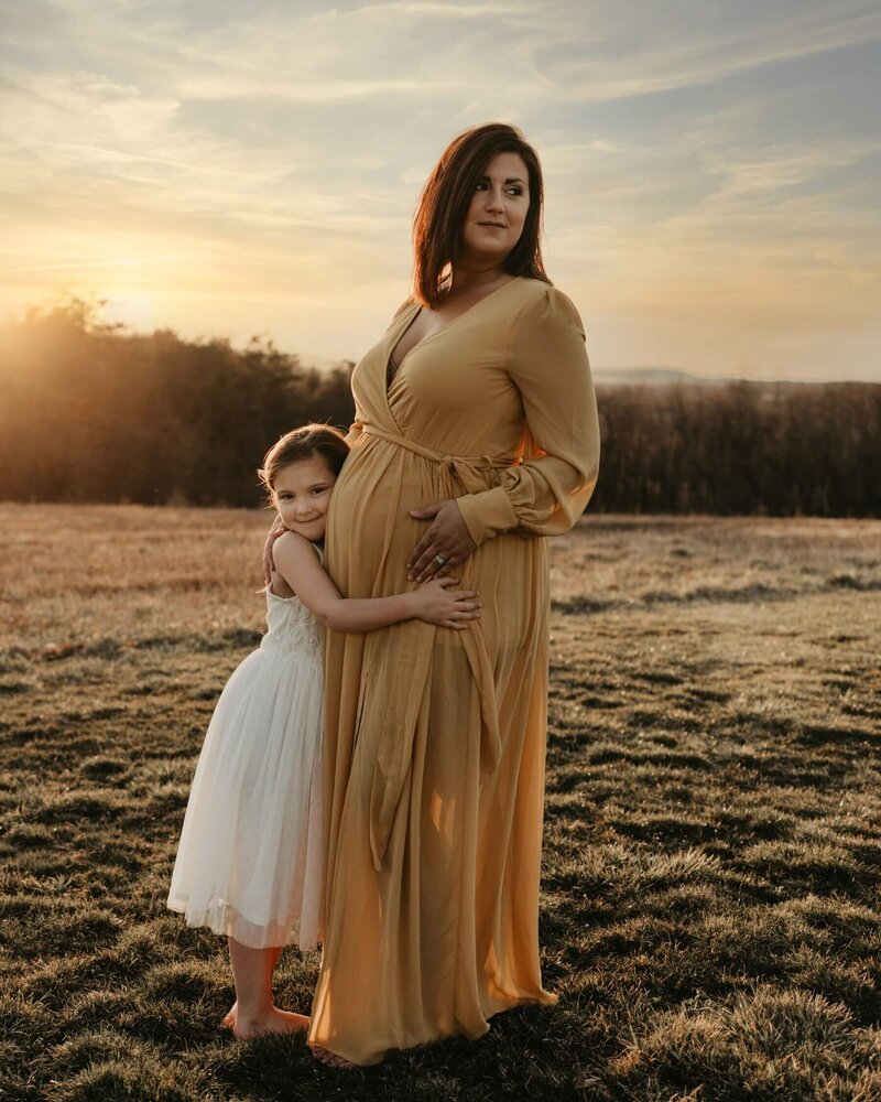 A pregnant woman and her daughter standing in a field at sunset, captured beautifully by a Pittsburgh maternity photographer.