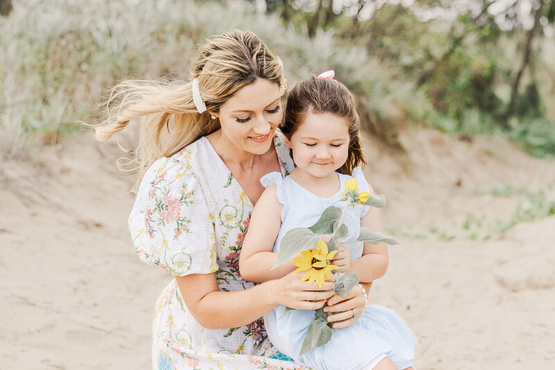 Mother with daughter sitting on her knee, looking at sunflowers at the beach. Captured by Yeppoon Photographer Julie-Anne Photography.
