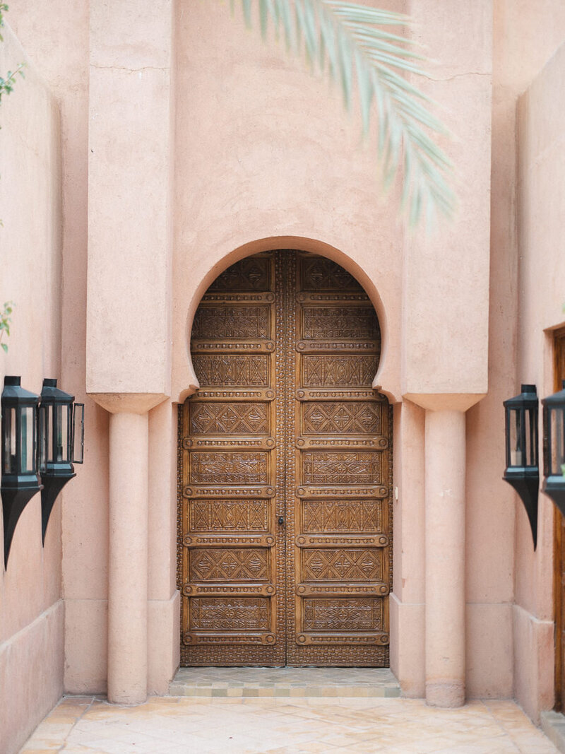 A moroccan handcrafted door stands behind beautiful teracotta coloured moroccan walls, taken at Amanjena in Marrakech by destination wedding phtographer Abul