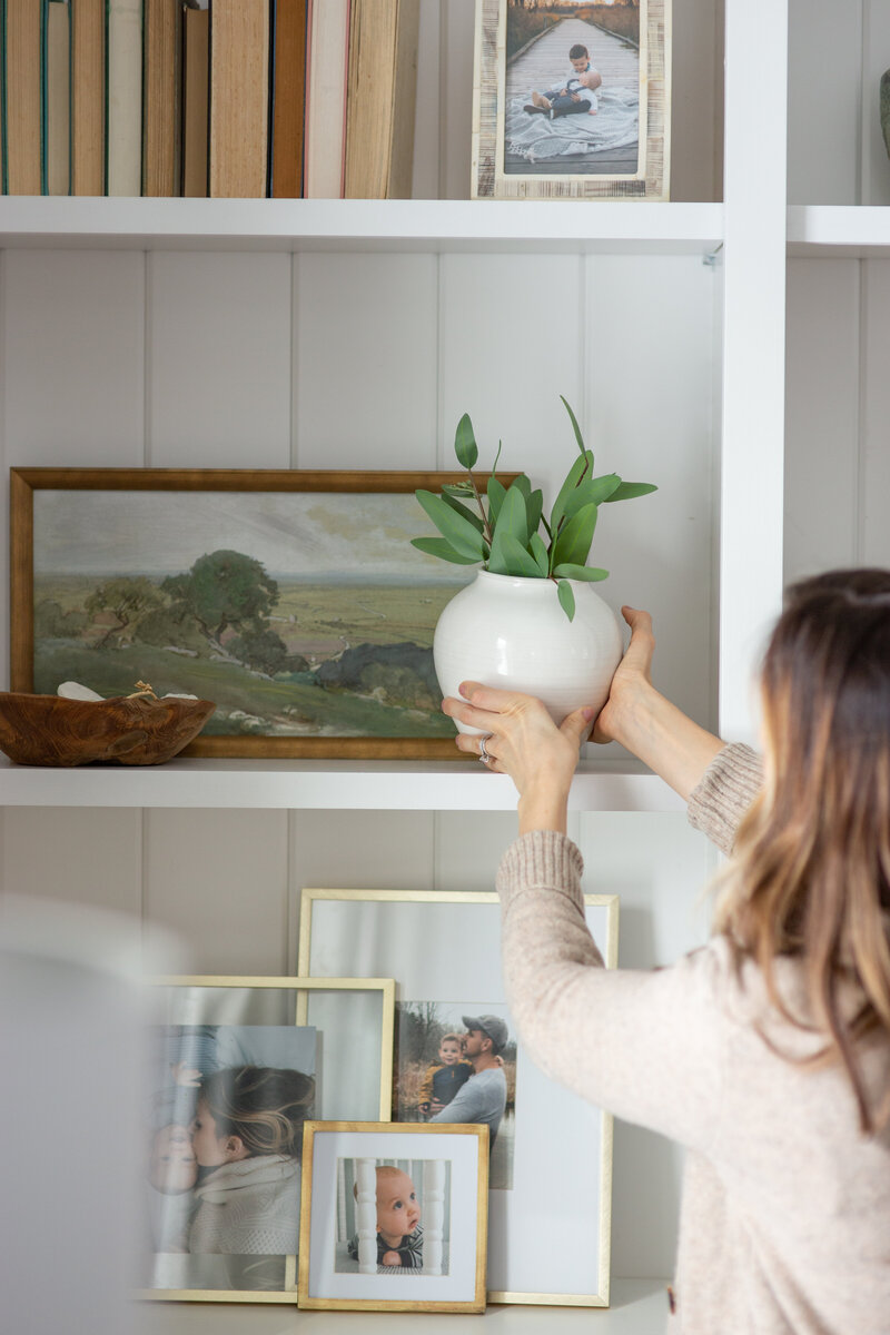 Woman placing a potted plant on a shelf that has been styled with photos and books