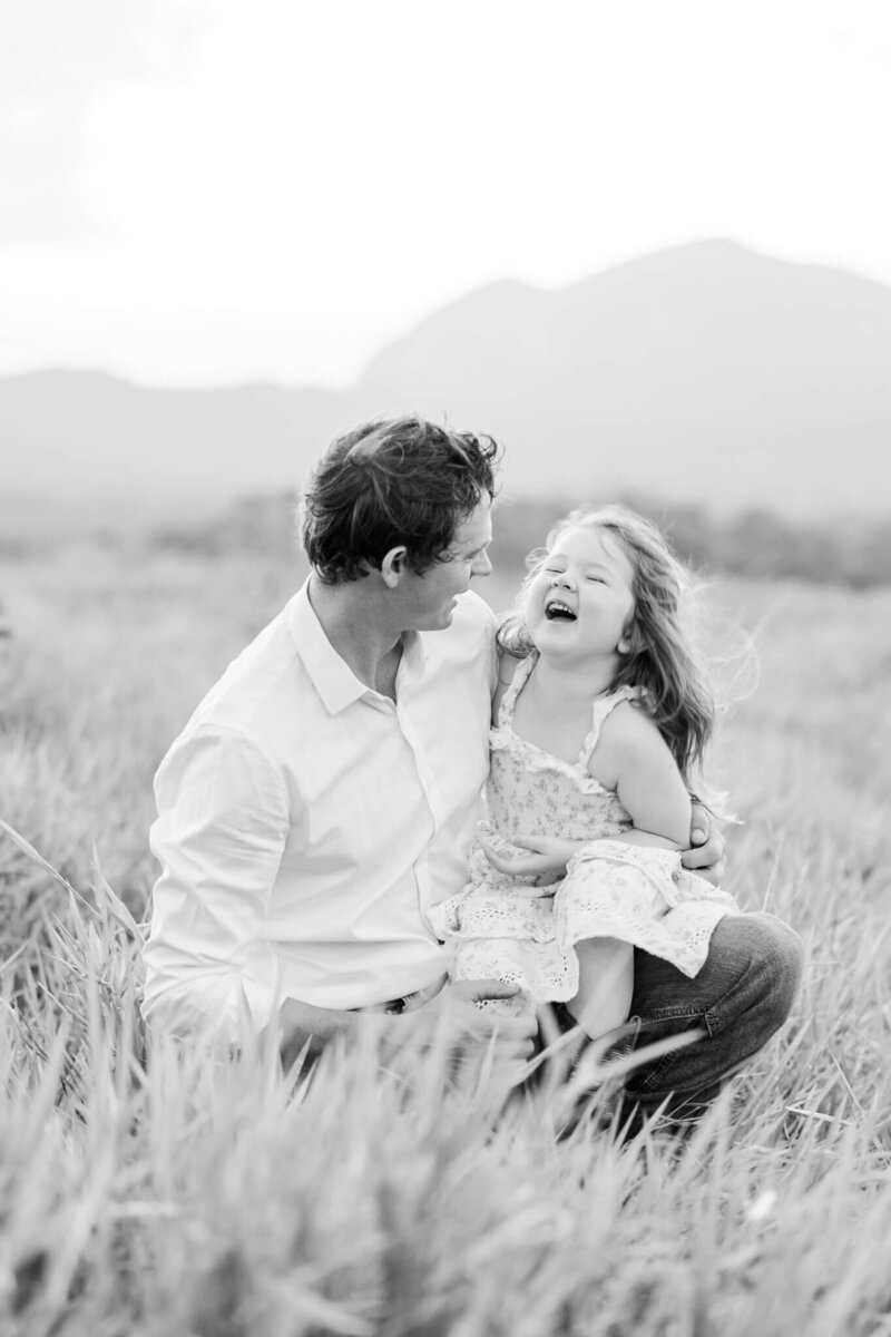 Black and white image of a little girl laughing happily up at her dad while sitting on his lap as he kneels in a grassy field.