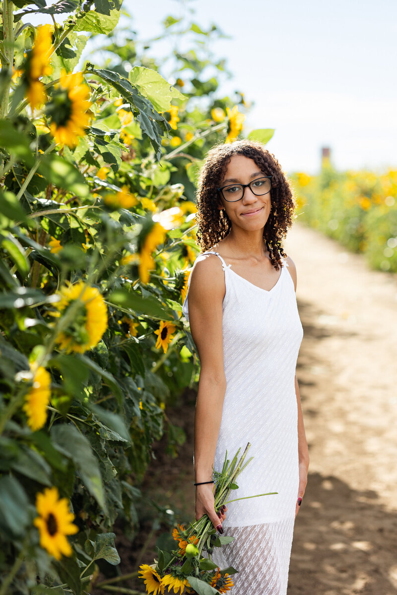 A young woman in a white lace dress stands in a sunflower field holding a bouquet of sunflowers and smiles at the camera.