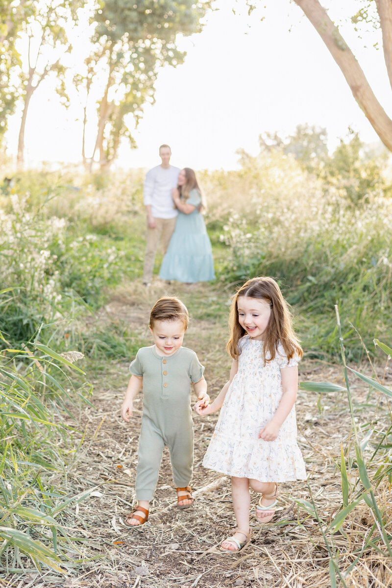 A young brother and sister hold hands while walking down a nature path, with their parents embracing in the blurred background.
