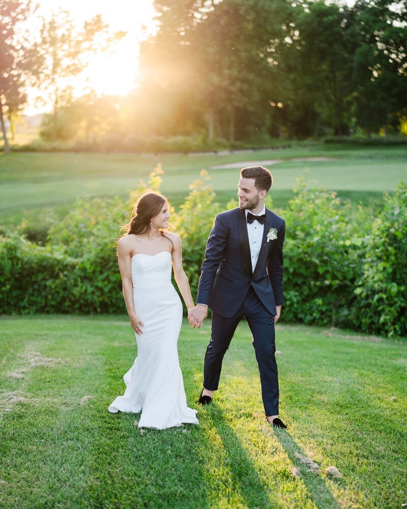A bride and groom hold hands and smile at each other with the golden sun setting behind them