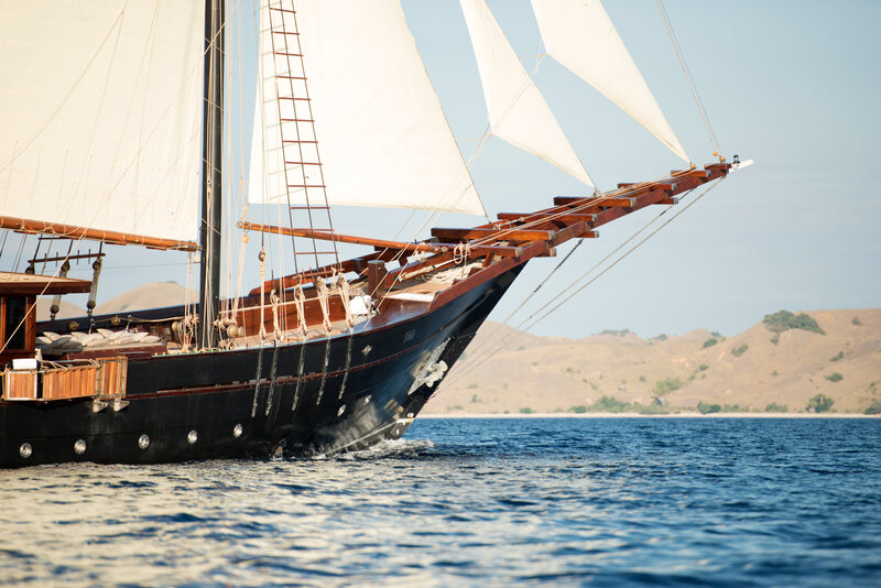A majestic wooden sailing yacht glides through the turquoise waters of Indonesia, with rugged island landscapes in the background.