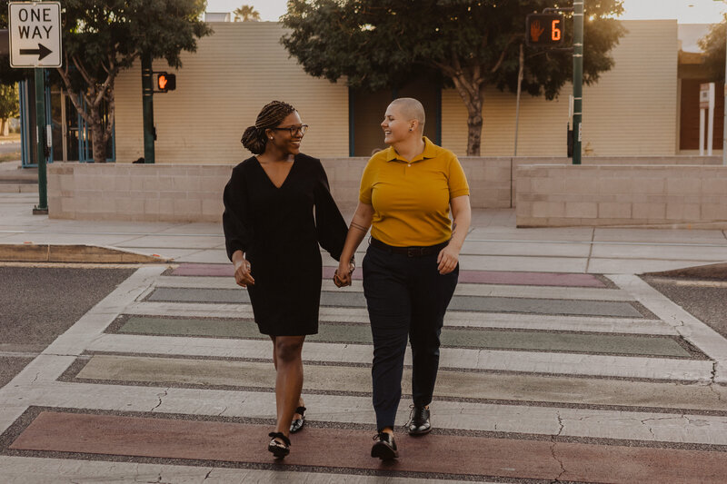 A queer couple holding hands as they cross the street.