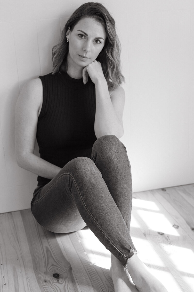 A minimal studio look - a woman in jeans sits in a soft pool of light casually glancing up at the camera