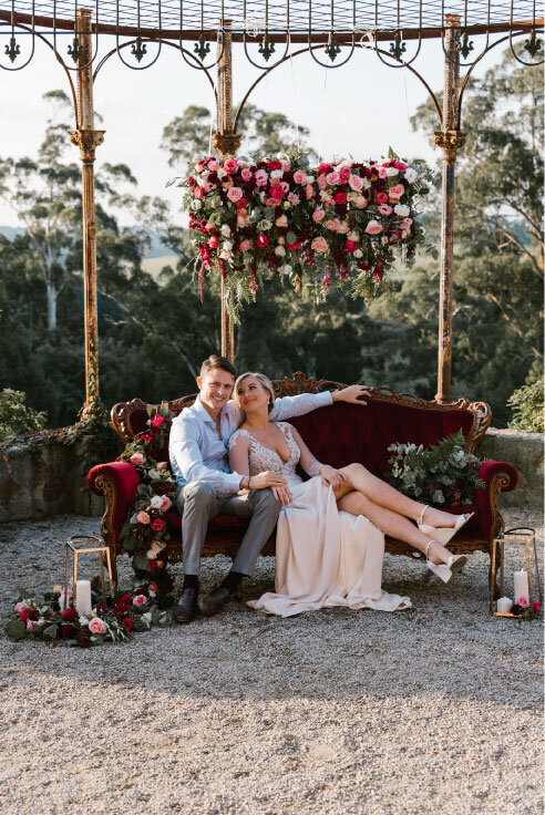 Bride and groom sitting on red velvet couch with floral chandelier in red and pink