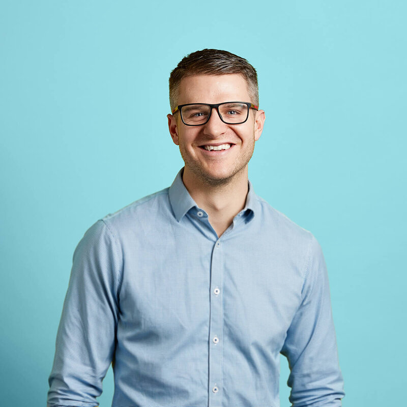 Smiling man with glasses wearing a blue checked shirt against a blue background.