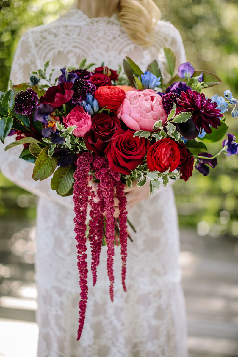 A bride in an empire neck-line lace wedding dress holds a flowing bouquet of large, vibrant red, pink, burgundy, purple, and blue flowers.