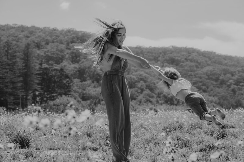 A woman is twirling a child by the arms in a field with trees and hills in the background. The image is in black and white.