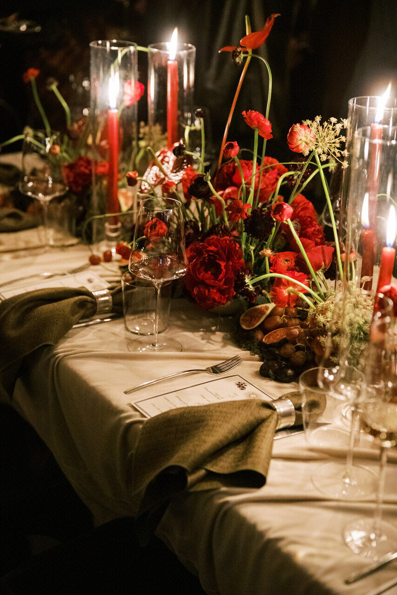 Dramatic wedding tablescape with deep red florals, tall candles, and elegant tableware creating a romantic and moody ambiance.