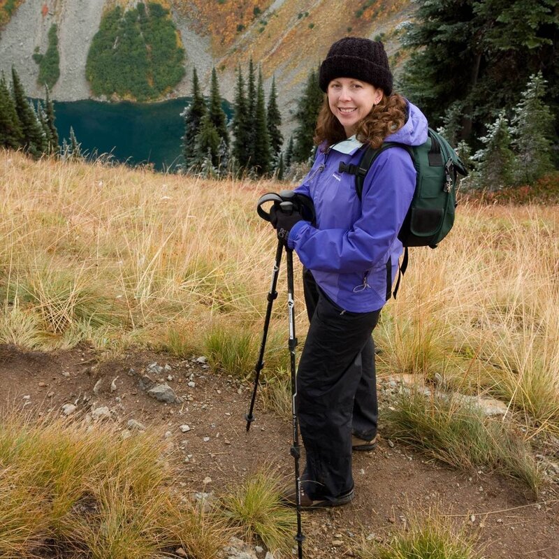 A woman stands on a hiking trail, smiling at the camera. She is wearing a purple jacket, black pants, a black beanie, and a green backpack. She is holding hiking poles, and there is a scenic view of a lake and mountains in the background.