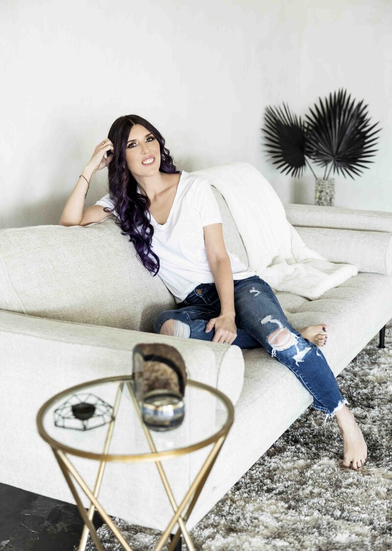 Laura sits on a white couch and smiles at the camera, resting her arm against the top of the couch and leaning against her hand. She is wearing a white v-neck t-shirt with ripped skinny jeans. Her feet are bare, painted purple toes visible. A black fanned plant is visible in the background against a white wall.